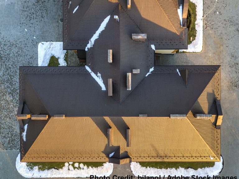 An aerial view shows a dark metal roof with multiple ridgelines and valleys, dusted with patches of melting snow.