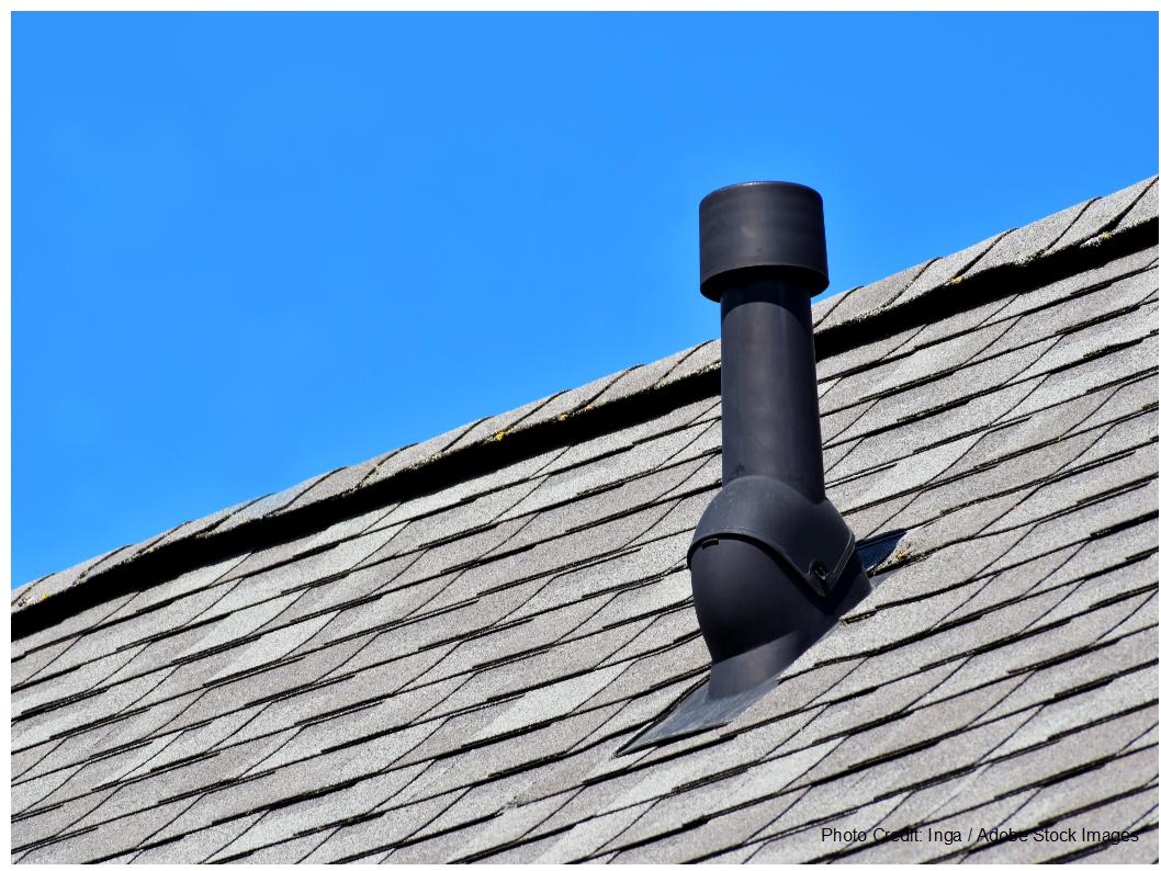 A black plumbing vent pipe rises from a gray shingle roof beneath a clear blue sky.