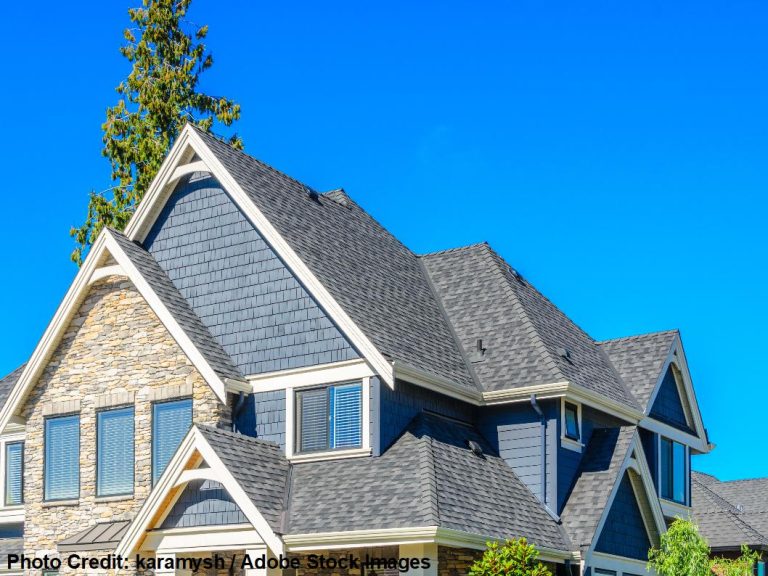A two-story home with stone accents and dark siding features a multi-gable dimensional shingle roof beneath a bright blue sky.