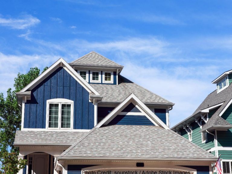 A blue two-story home with multiple gables features a gray dimensional shingle roof beneath a bright blue sky with light clouds.