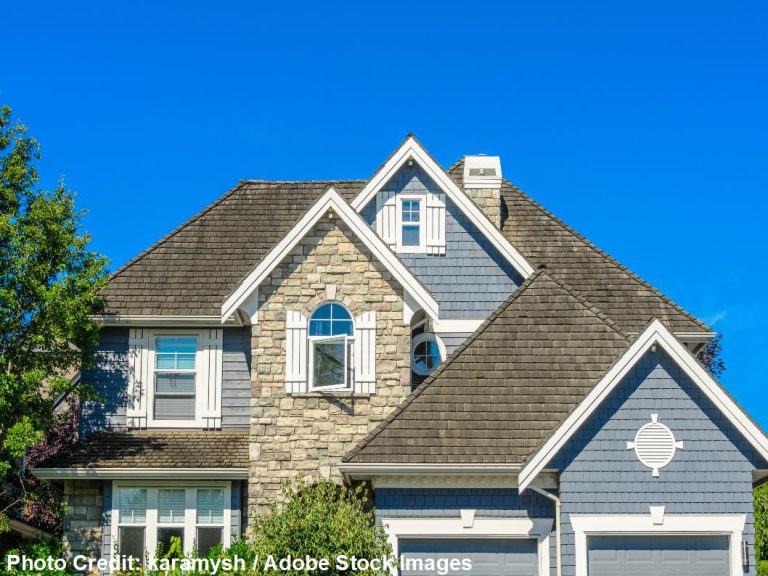 A blue-gray two-story home with stone accents features a multi-gable dimensional shingle roof beneath a bright blue sky.