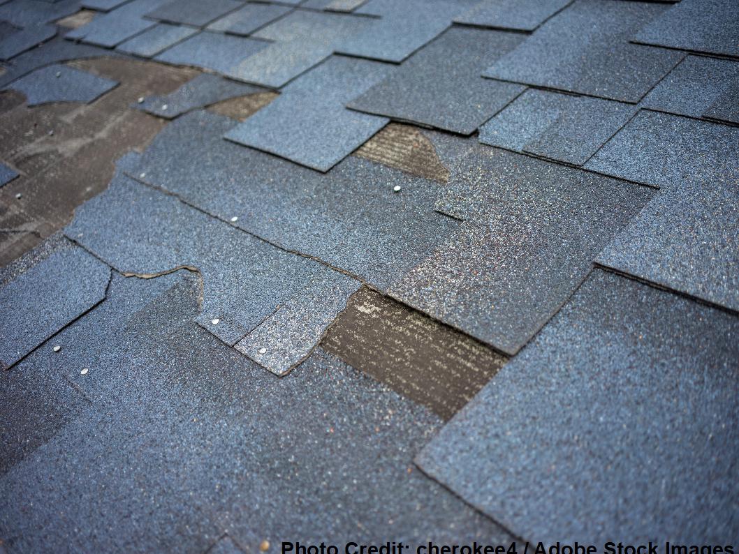 A close-up shows worn blue-gray asphalt shingles with visible granule loss and a patch of exposed dark underlayment.