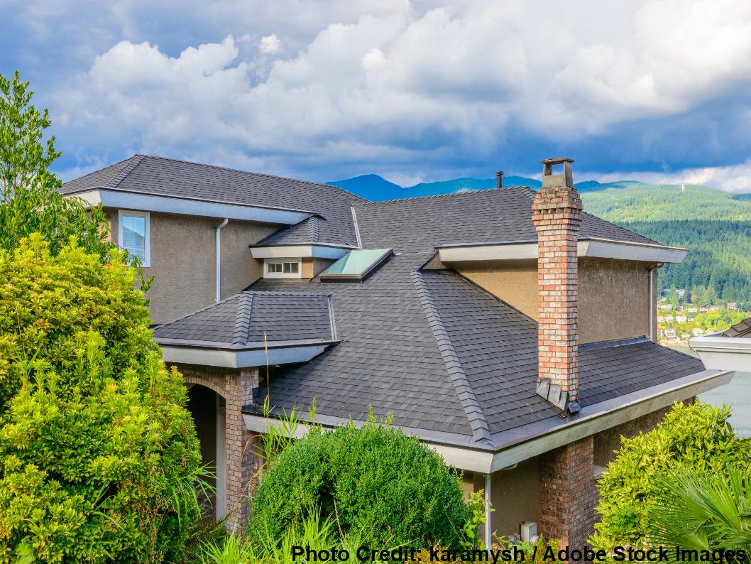 Elevated view of a large home with a dark gray shingle roof featuring multiple hips, valleys, dormers, and a brick chimney.