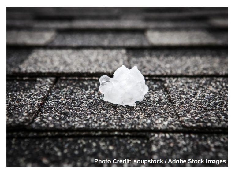 A jagged chunk of hail sits on gray asphalt shingles, illustrating the kind of impact damage severe storms can leave behind.