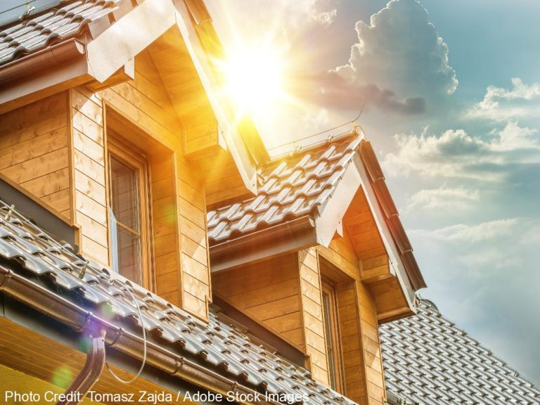 Bright sunlight flares over a wooden dormer and tile roof beneath a partly cloudy blue sky.