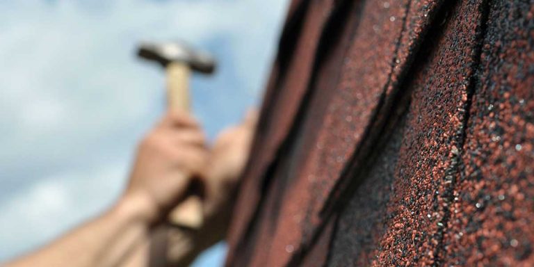 A close-up of blurred hands holding a hammer beside the sharp textured edge of reddish-brown roof shingles.