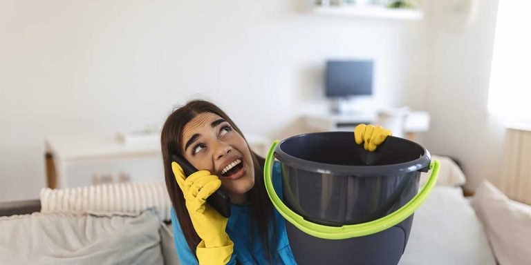 A worried woman wearing rubber gloves holds a bucket under a ceiling leak while talking on the phone.