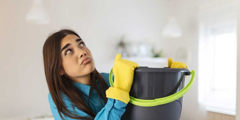 Frustrated woman wearing yellow gloves holds a bucket indoors while looking up at a leaking ceiling needing roof replacement.